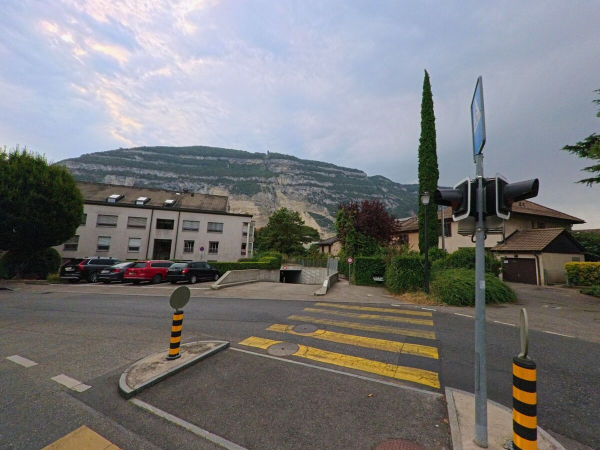 In Veyrier (1255, Kanton Genf) fuehrt ein Fussgaengerueberweg mit Verkehrsinsel ueber die Strasse, im Hintergrund sind Wohnhaeuser und der markante Berghang zu sehen.