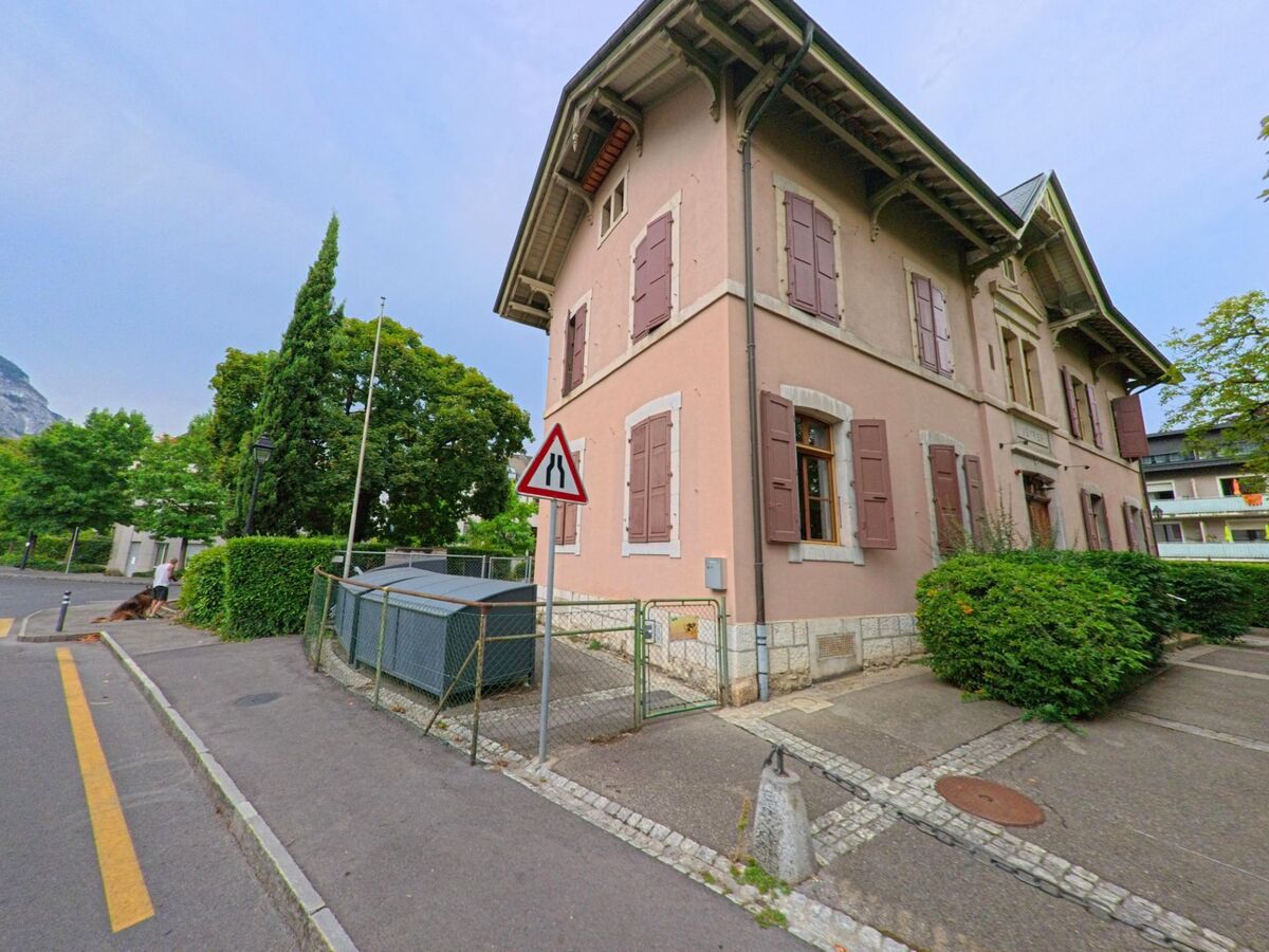 In Veyrier (1255, Kanton Genf) steht ein rosa gestrichenes Haus mit geschlossenen Fensterlaeden an einer Strasse mit Gehweg und Strassenzeichen.
