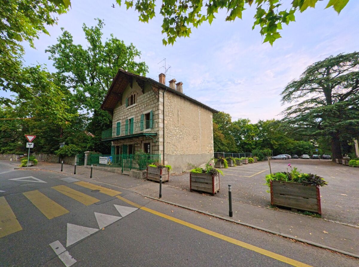 In Veyrier (1255, Kanton Genf) steht ein Steinhaus mit gruenen Fensterlaeden neben einem Parkplatz, umgeben von grossen Baeumen.