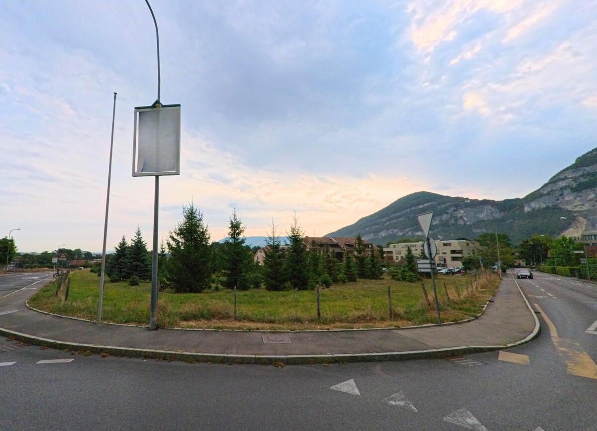 In Veyrier (1255, Kanton Genf) fuehrt eine Strasse an Wiesen und jungen Nadelbaeumen vorbei, im Hintergrund erheben sich bewaldete Berghaenge.
