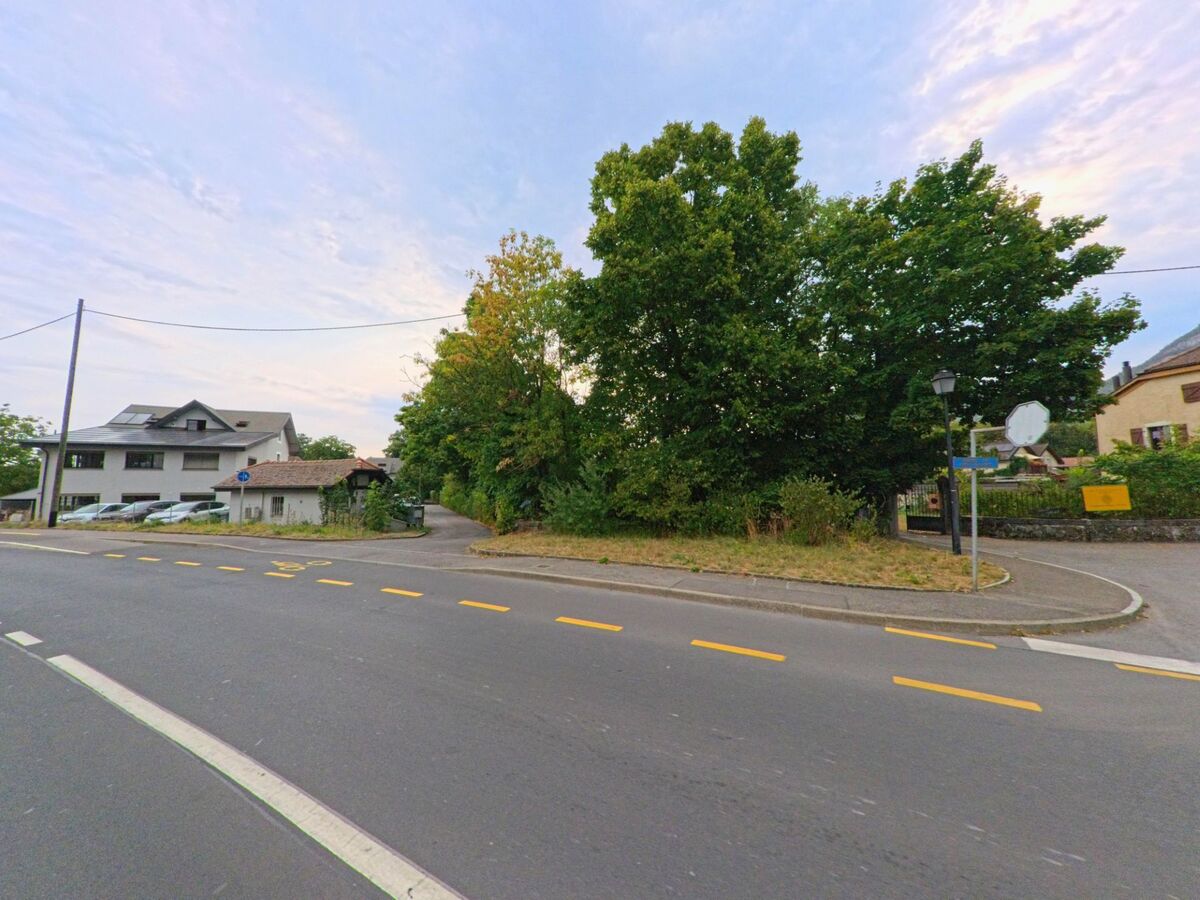 An einer Strassenecke in Veyrier (1255, Kanton Genf) zweigt ein schmaler Weg zwischen Wohnhaeusern und Baeumen ab.