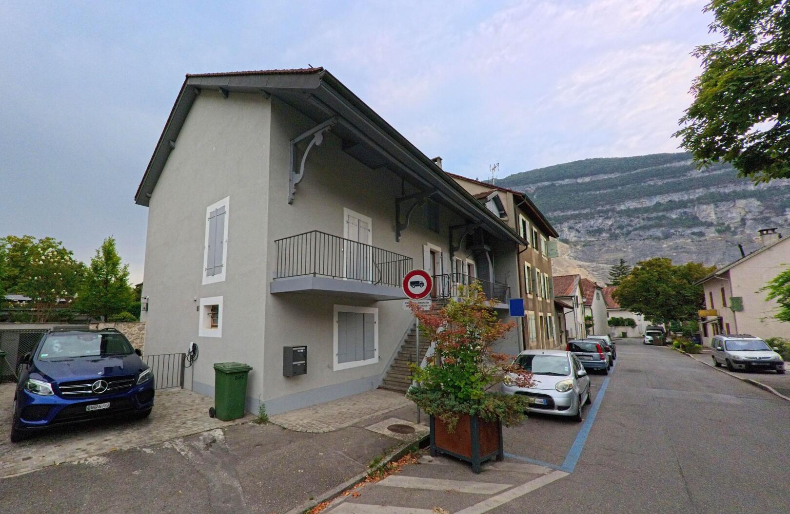 In Veyrier (1255, Kanton Genf) reihen sich Wohnhaeuser mit hellen Fassaden entlang einer schmalen Strasse, im Hintergrund ist der steile Berghang zu erkennen.