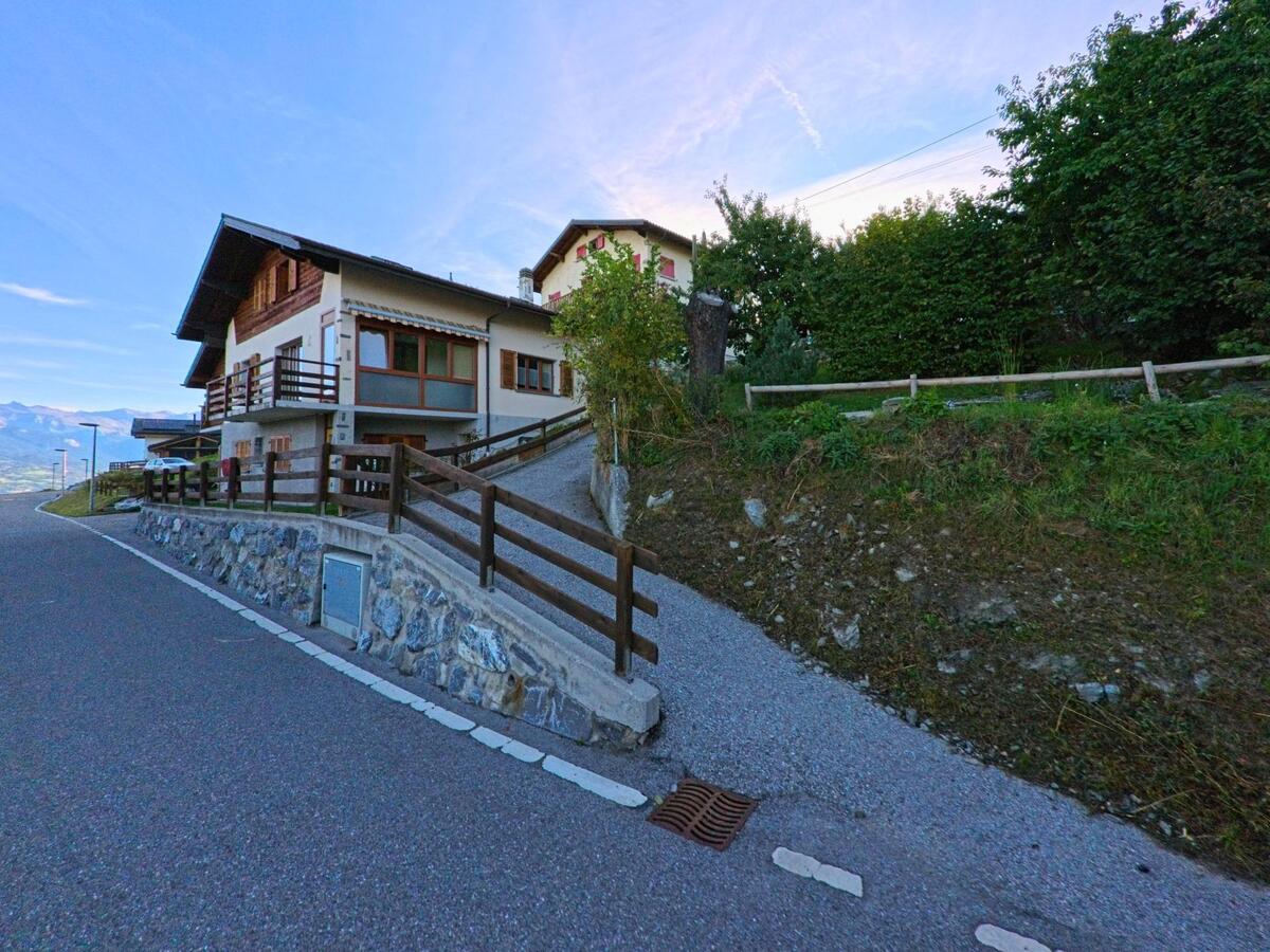 Ein Wohnhaus mit Holzbalkonen in Veysonnaz (1993, Kanton Wallis) steht an einer schmalen Strasse mit ansteigendem Weg und Natursteinmauer.