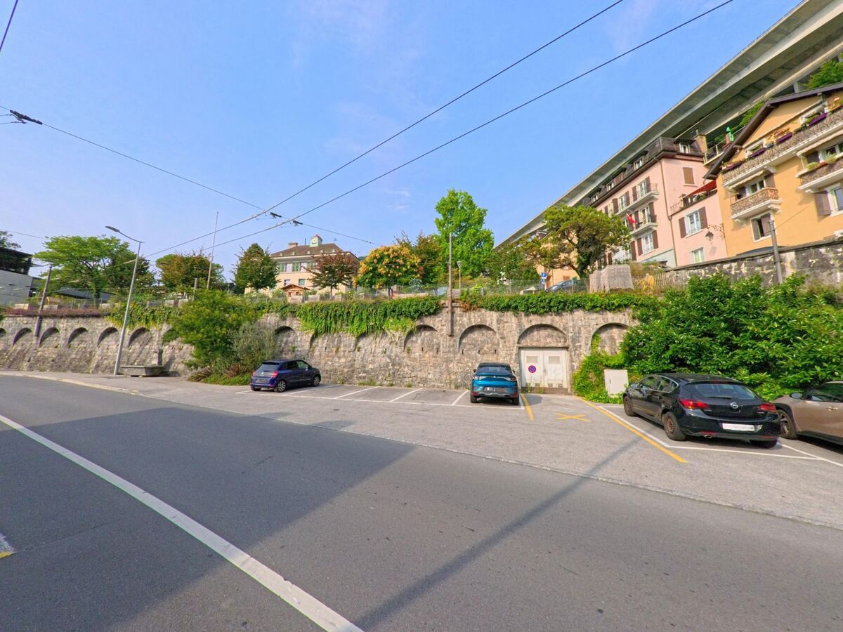 In Veytaux (1820, Kanton Waadt) liegt ein Parkplatz unterhalb einer alten Steinmauer mit begrünten Bögen und dahinterliegenden Wohnhäusern.