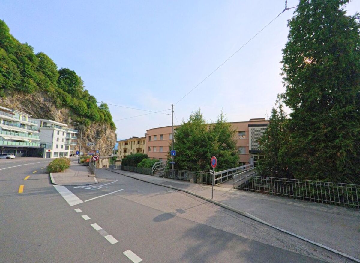 Eine ruhige Strasse in Veytaux (1820, Kanton Waadt) führt zwischen modernen Gebäuden und einer Felswand entlang, flankiert von Bäumen und Gehwegen.