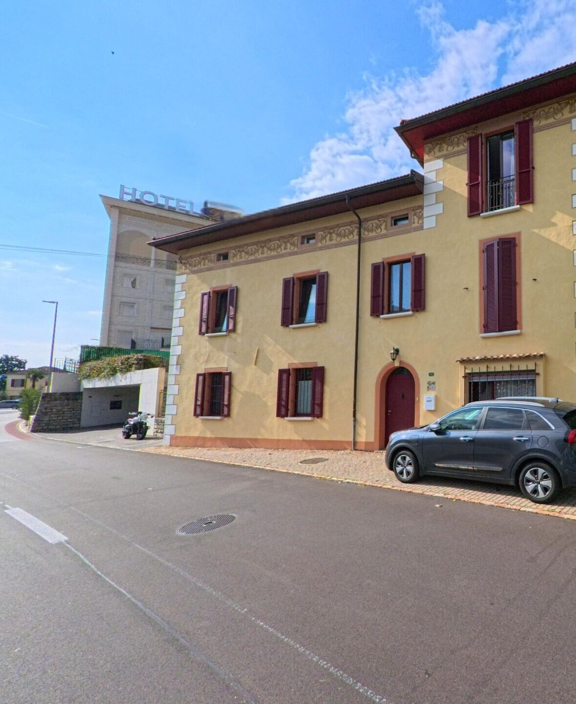 Entlang einer ruhigen Strasse stehen traditionell gestaltete Wohnhäuser in Vico Morcote (6921, Kanton Tessin), während im Hintergrund ein Hotelgebäude über die Dächer hinausragt.
