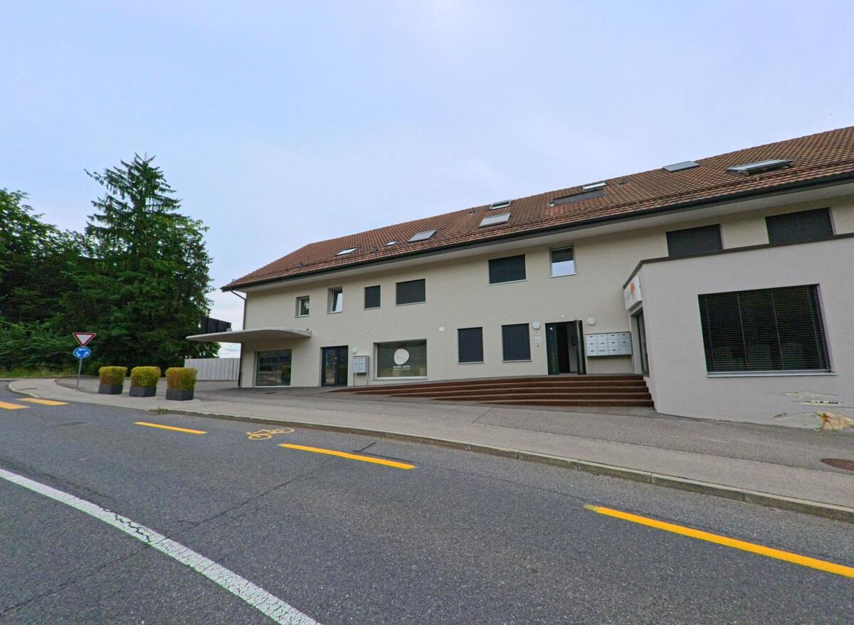 In Villars-sur-Glâne (1752, Kanton Freiburg) steht ein modernes Gewerbegebäude mit rotem Ziegeldach und breiter Treppenanlage direkt an einer leicht abschüssigen Strasse.