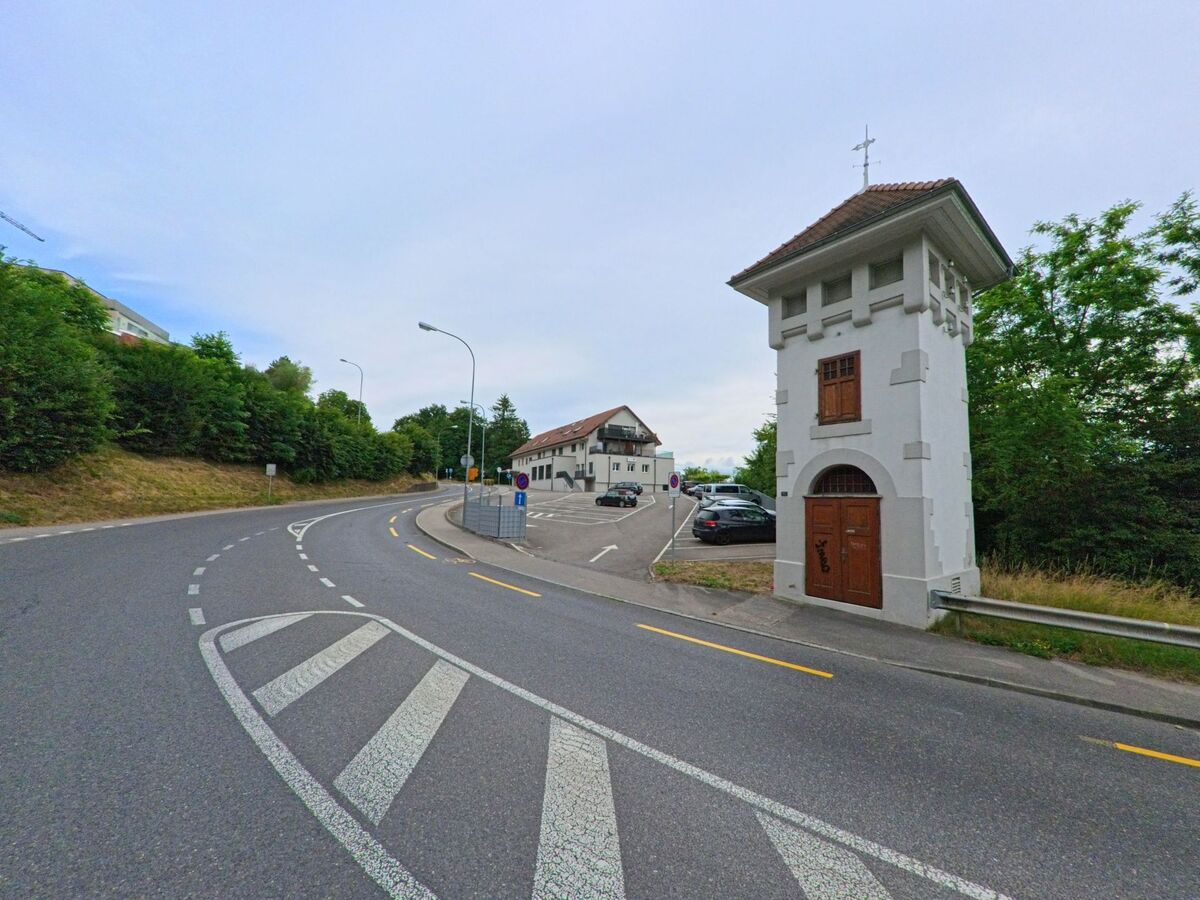In Villars-sur-Glâne (1752, Kanton Freiburg) steht ein historischer Turm mit hölzernem Eingang an einer stark befahrenen Strassenkurve neben einem Wohnquartier.