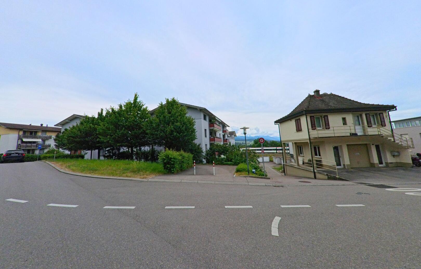 In Villars-sur-Glâne (1752, Kanton Freiburg) verzweigt sich eine Quartierstrasse zwischen einem älteren Wohnhaus mit Balkon und modernen Mehrfamilienhäusern im Hintergrund.