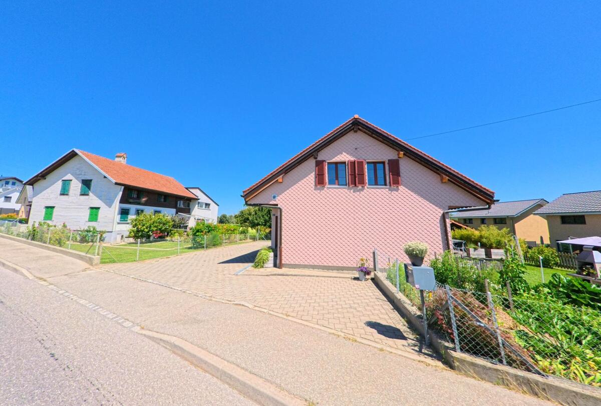 In einer Wohnsiedlung von Villaz (1690, Kanton Freiburg) steht ein rosa Einfamilienhaus mit Garten neben einem traditionellen Wohnhaus mit rotem Ziegeldach.
