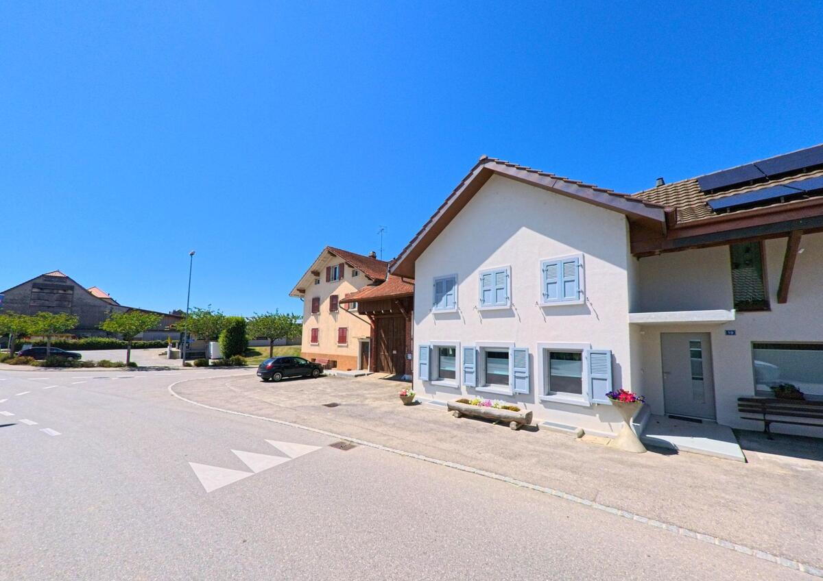 An einer breiten Kreuzung in Villaz (1690, Kanton Freiburg) stehen Wohnhäuser mit hellen Fassaden und Fensterläden, im Hintergrund sind Wirtschaftsgebäude zu sehen.