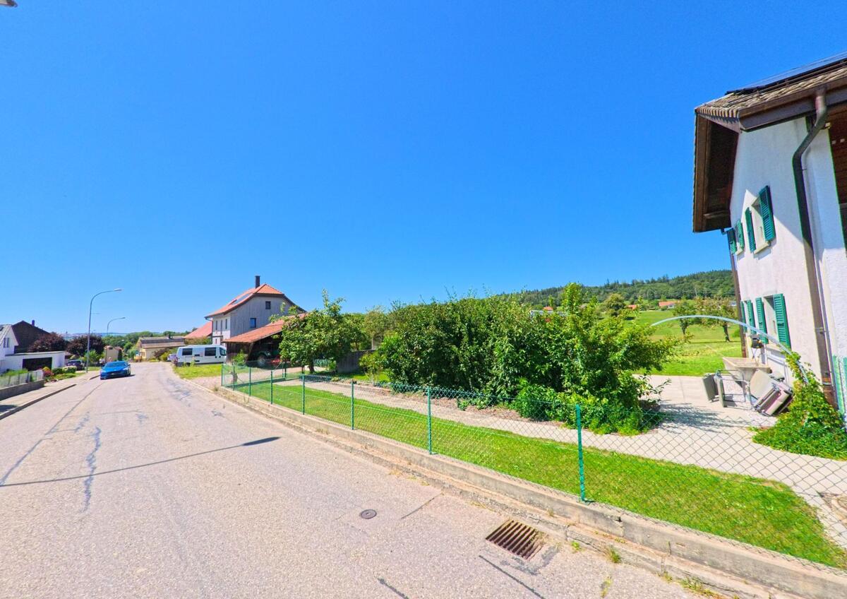 In einer Wohnstrasse von Villaz (1690, Kanton Freiburg) stehen Einfamilienhäuser mit Gärten, im Hintergrund sind Felder und Wälder sichtbar.