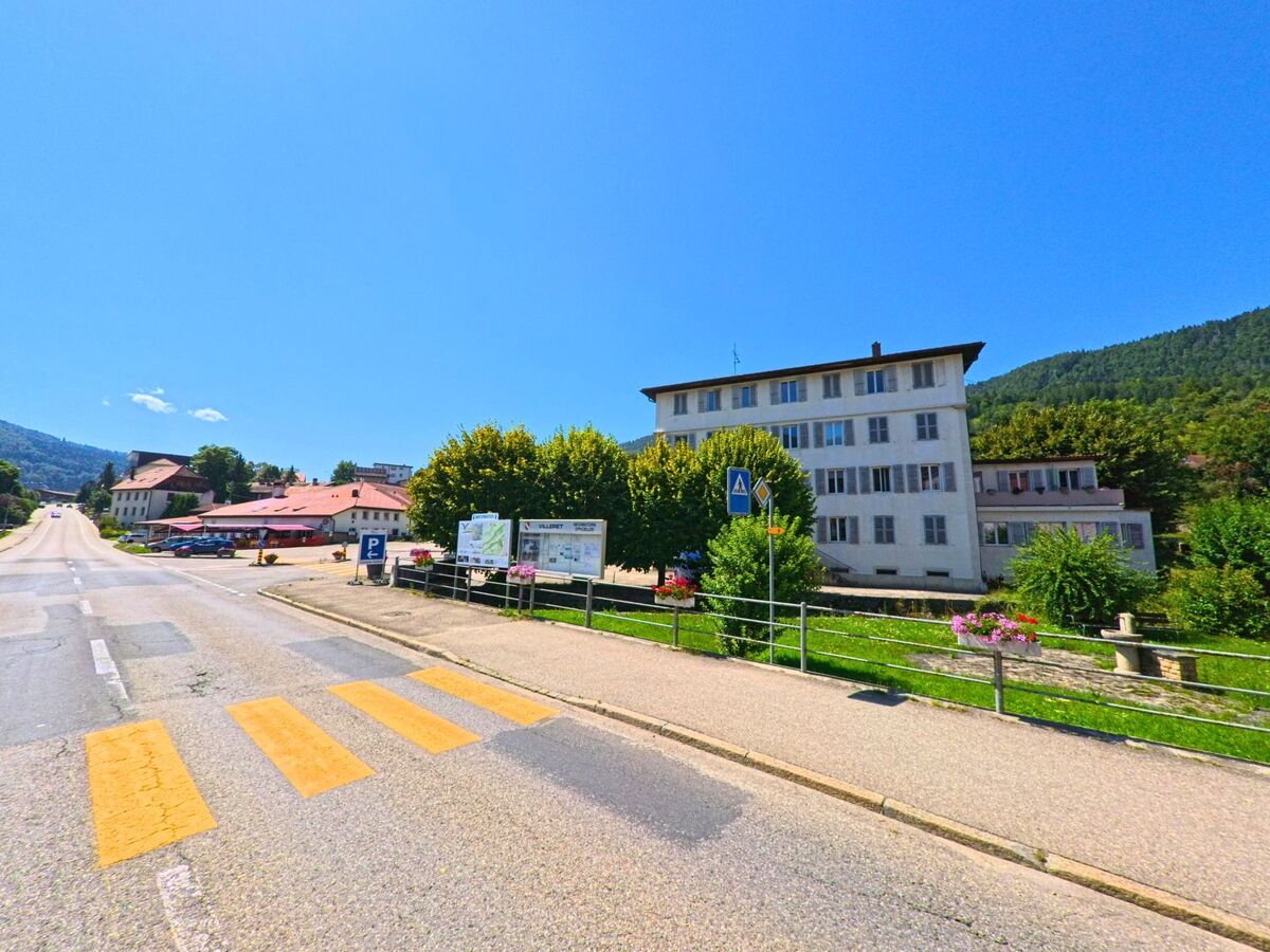 An einer breiten Strasse in Villeret (2613, Kanton Bern) steht ein grosses, weisses Gebaeude mit mehreren Stockwerken neben einem kleinen Park mit Blumen.