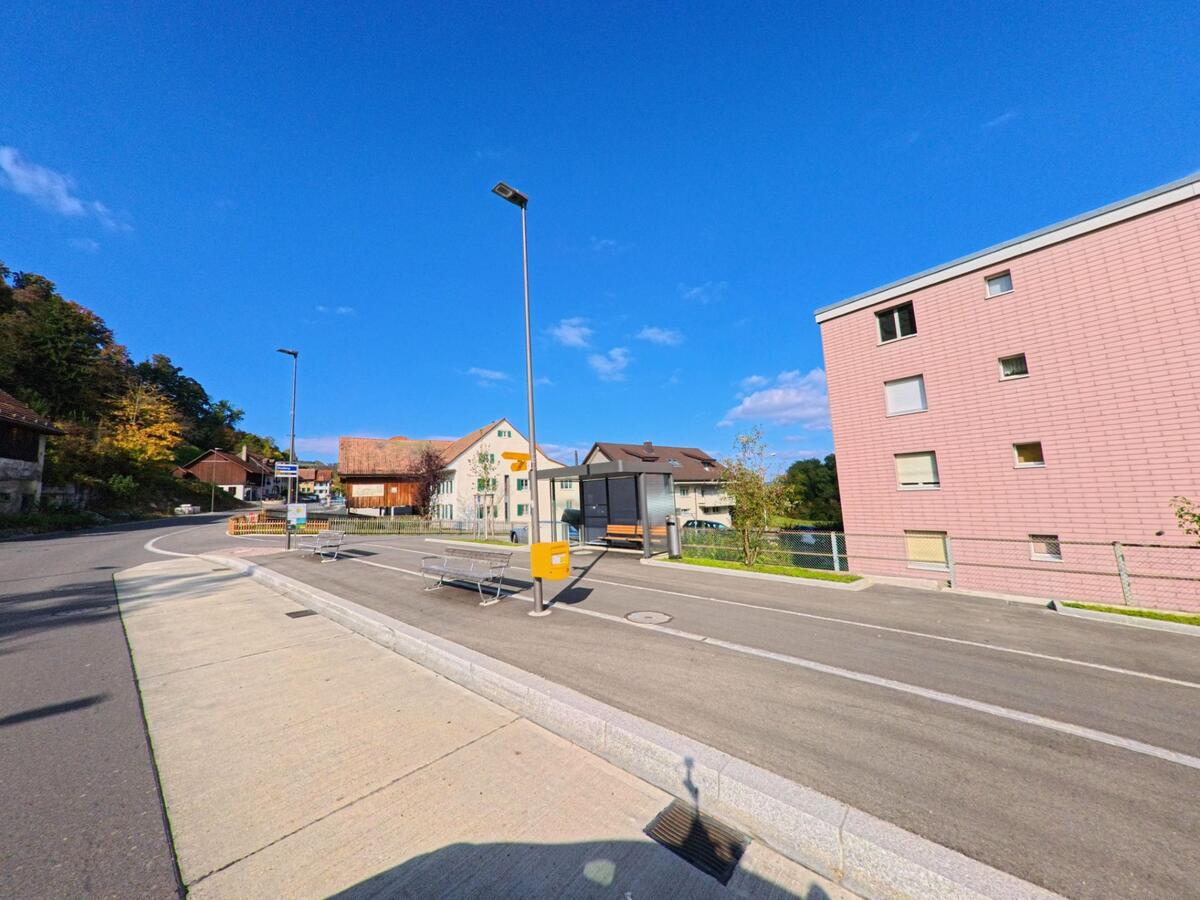 An einer Bushaltestelle in Villigen (5233, Kanton Aargau) stehen Sitzbaenke und ein gelber Briefkasten vor Wohnhaeusern, waehrend sich die Strasse sanft durch das Quartier windet.