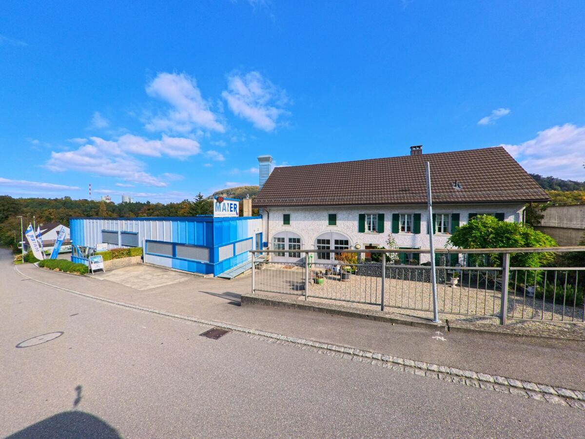 In Villigen (5233, Kanton Aargau) stehen ein modernes blau-weisses Gewerbegebaeude und ein traditionelles Wohnhaus mit gruenen Fensterlaeden nebeneinander an einer leicht abfallenden Strasse.