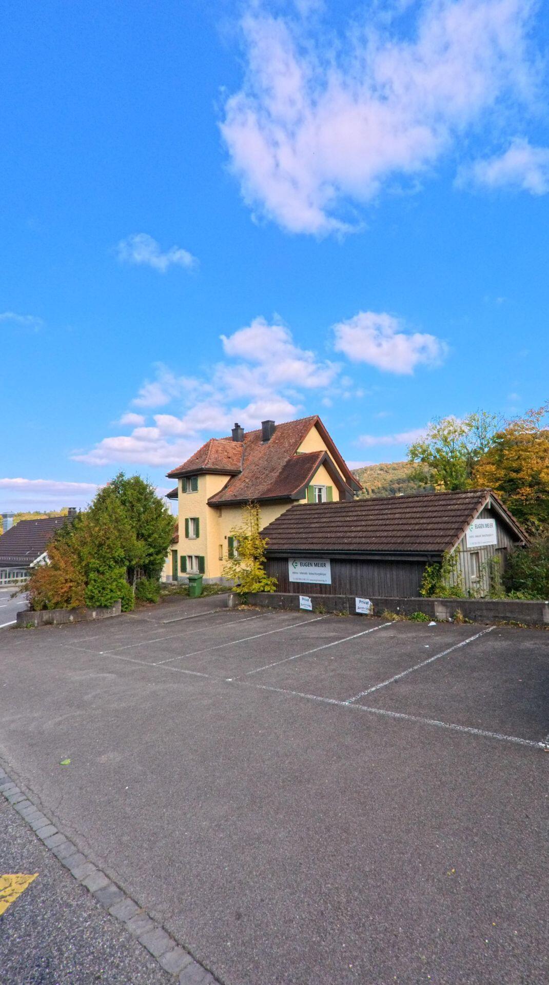 Ein kleiner Parkplatz in Villigen (5233, Kanton Aargau) liegt vor einem gelben Wohnhaus und einem Holzschuppen, die von herbstlicher Vegetation umgeben sind.