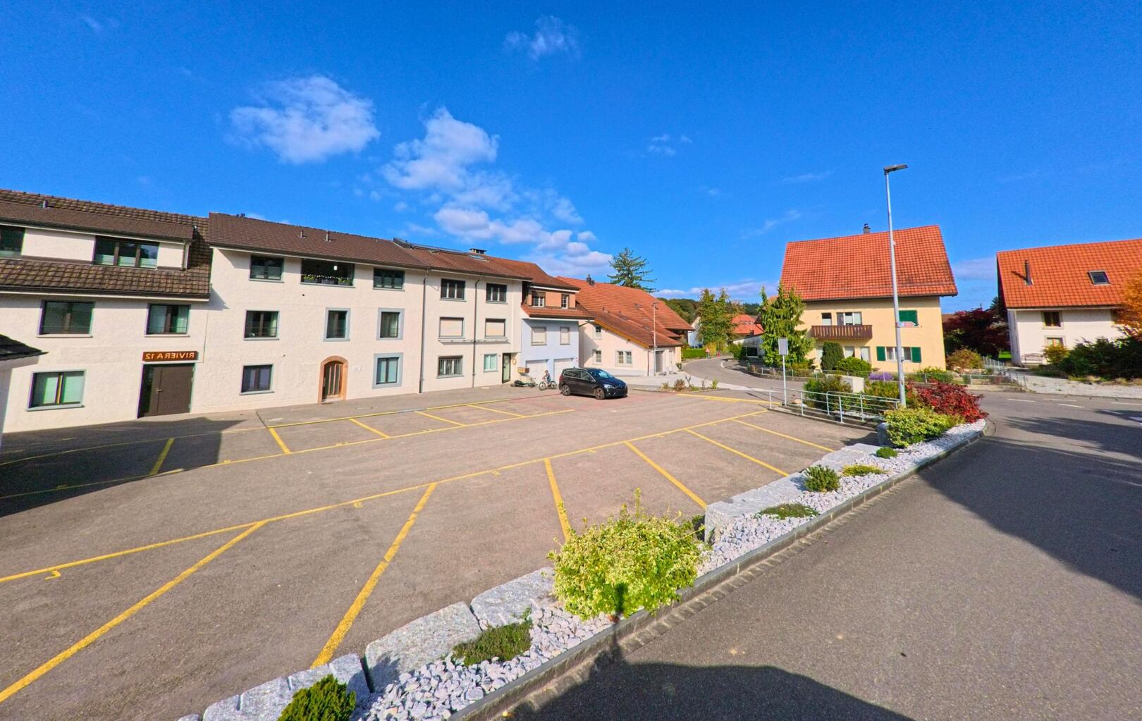 Ein nahezu leerer Parkplatz in Villigen (5233, Kanton Aargau) liegt zwischen Wohnhaeusern mit roten Daechern und einem kleinen Gartenbereich im Vordergrund.