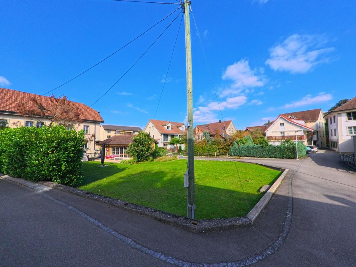 Zwischen mehreren Wohnhaeusern in Villigen (5233, Kanton Aargau) liegt eine gruene Rasenflaeche, die von Wegen und einem zentralen Strommast eingerahmt wird.