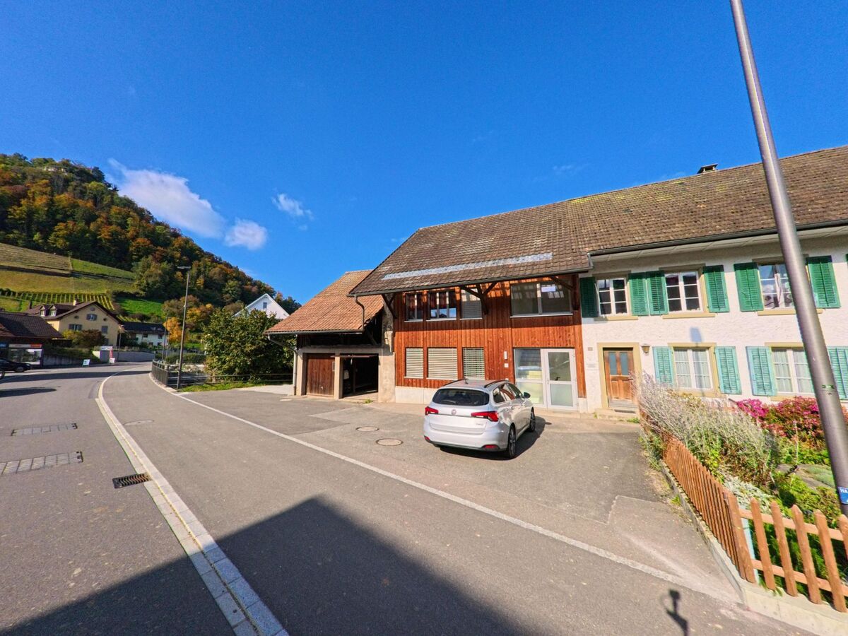In einer ruhigen Strasse von Villigen (5233, Kanton Aargau) stehen traditionelle Wohnhaeuser mit gruene Fensterlaeden vor einem herbstlich bewaldeten Hang.