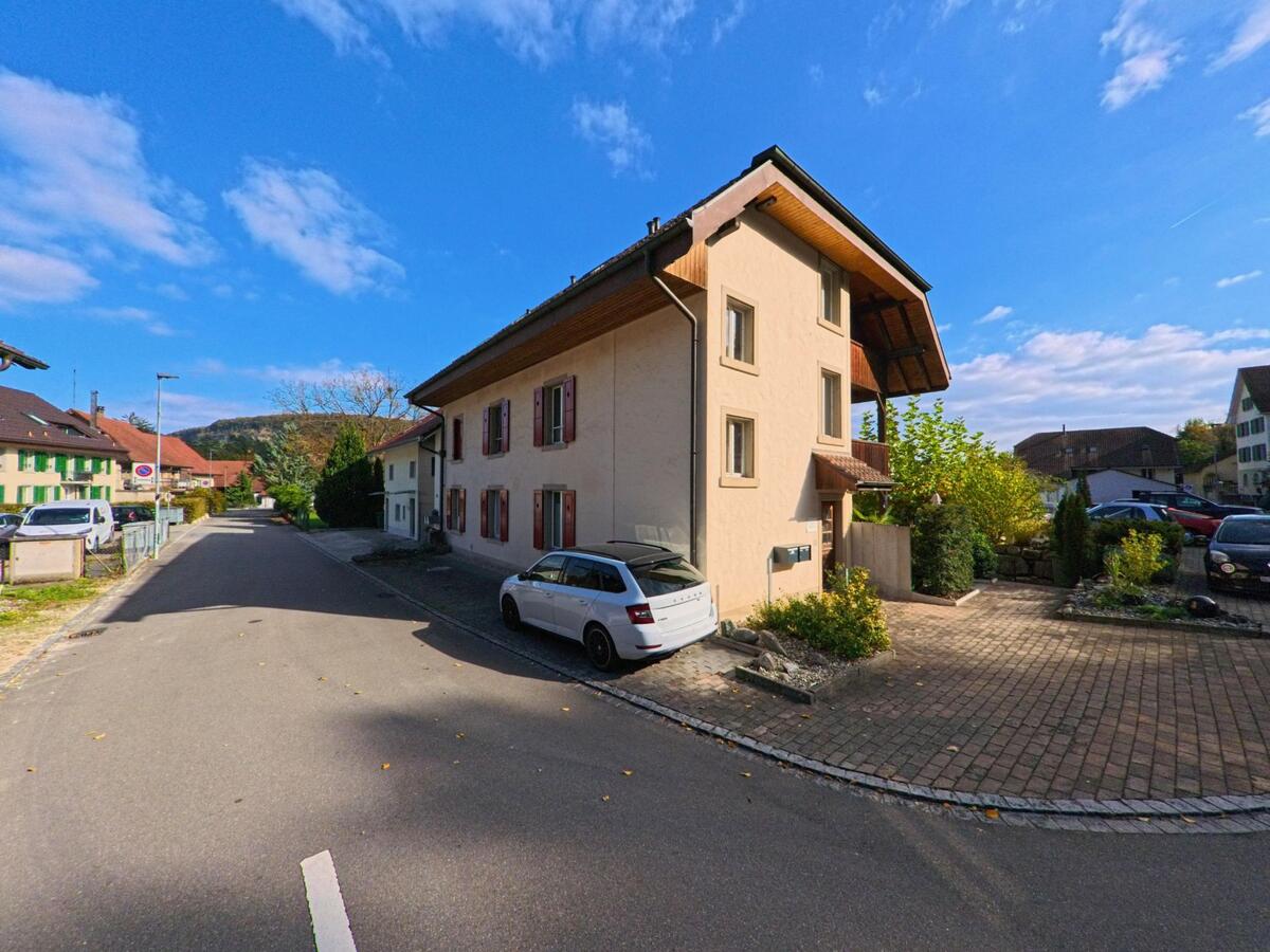In einer ruhigen Wohnstrasse von Villigen (5233, Kanton Aargau) stehen helle Haeuser mit Fensterlaeden entlang gepflegter Vorgaerten, waehrend im Hintergrund bewaldete Huegel sichtbar sind.