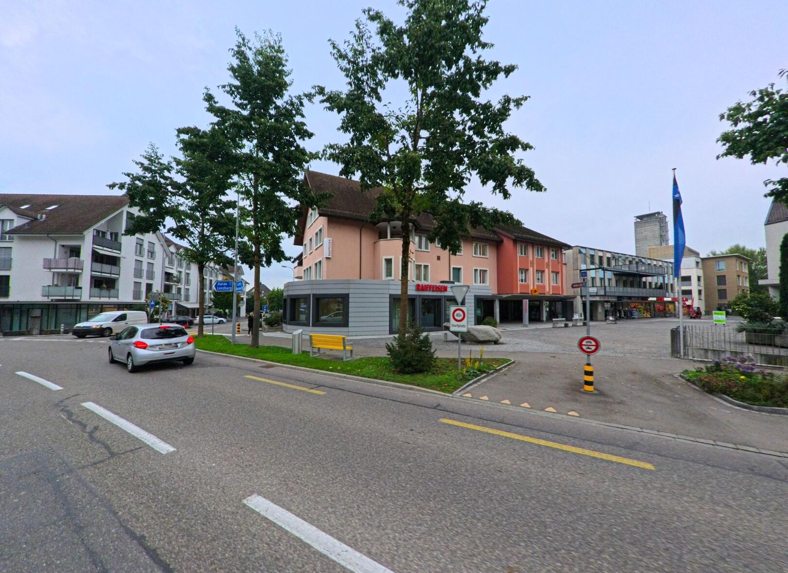 Im Zentrum von Villmergen (5612, Kanton Aargau) saeumen moderne Wohnhaeuser und ein rosa Gebaeude mit Bankfiliale den verkehrsreichen Strassenbereich.