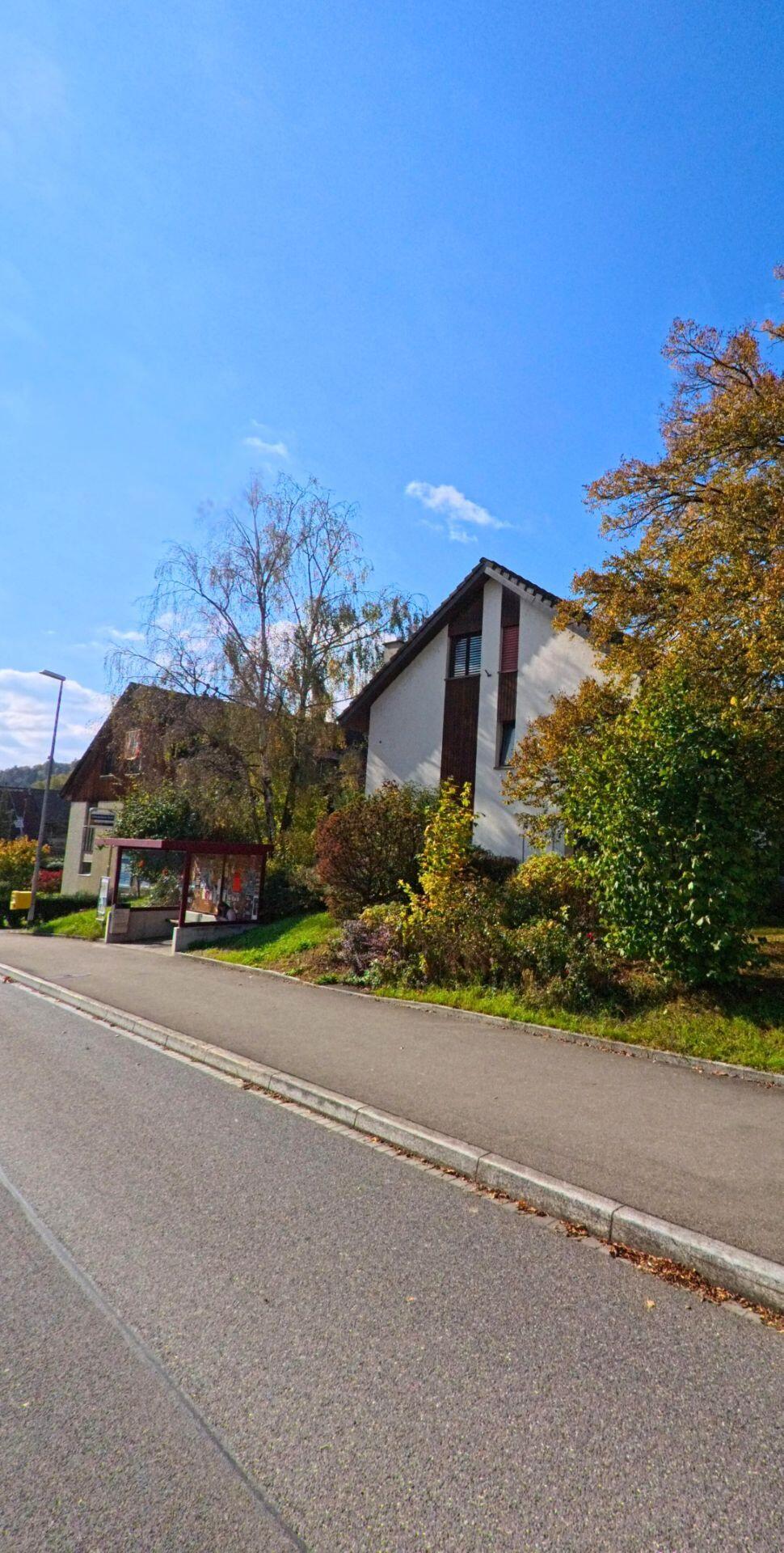 An einer Strasse in Villnachern (5213, Kanton Aargau) steht ein Wohnhaus neben einer kleinen Bushaltestelle, die von herbstlich gefaerbten Straeuchern und Baumen umgeben ist.
