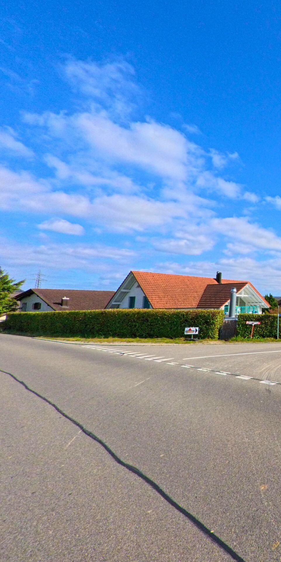 An einer Kreuzung in Villnachern (5213, Kanton Aargau) stehen Einfamilienhaeuser mit roten Daechern hinter einer dichten Hecke, waehrend sich der Himmel weit ueber der Strasse ausbreitet.