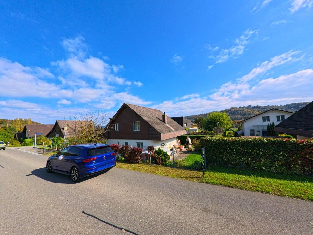 In einem Wohnquartier von Villnachern (5213, Kanton Aargau) saeumen Einfamilienhaeuser mit gepflegten Gaerten eine ruhige Strasse, vor der ein blaues Auto vorbeifaehrt und die bewaldeten Huegel im Hintergrund sichtbar sind.