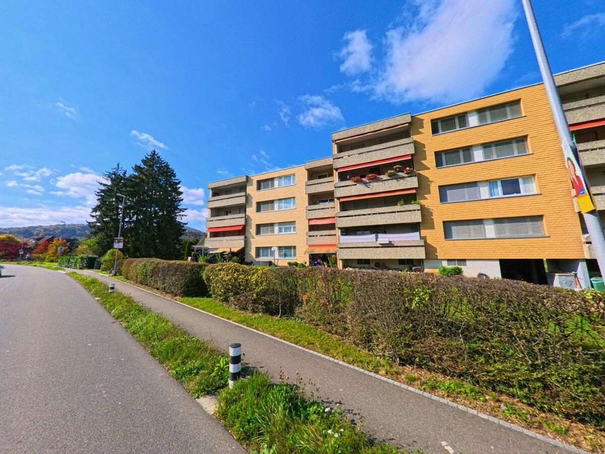 Entlang einer sanft geschwungenen Strasse in Villnachern (5213, Kanton Aargau) stehen mehrstöckige Wohnbauten mit Balkonen, die von niedrigen Hecken und einem separaten Gehweg eingefasst werden.