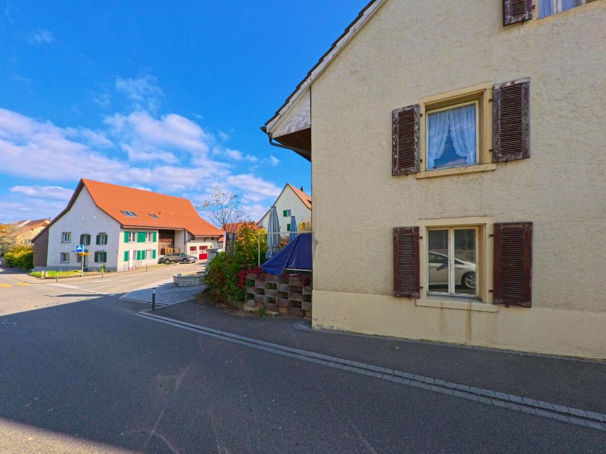 In einem ruhig wirkenden Quartier von Villnachern (5213, Kanton Aargau) stehen traditionelle Wohnhaeuser mit Fensterlaeden entlang einer kleinen Strasse, die zu weiteren Gebaeuden mit roten Daechern fuehrt.