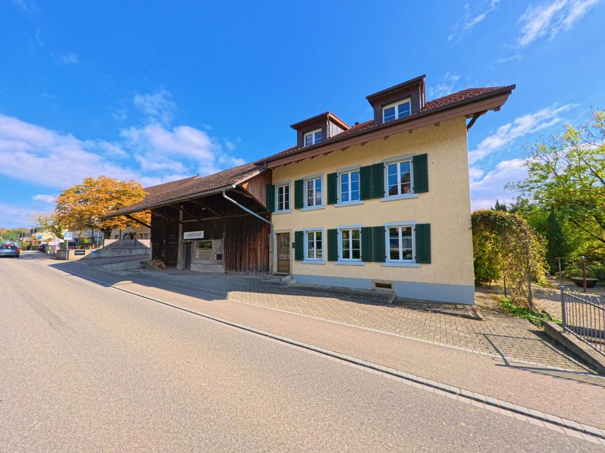Entlang einer sonnigen Strasse in Villnachern (5213, Kanton Aargau) steht ein traditionelles Gebaeude mit gruenen Fensterlaeden neben einem alten Holzschopf, waehrend herbstlich gefaerbte Baume die Szenerie begleiten.