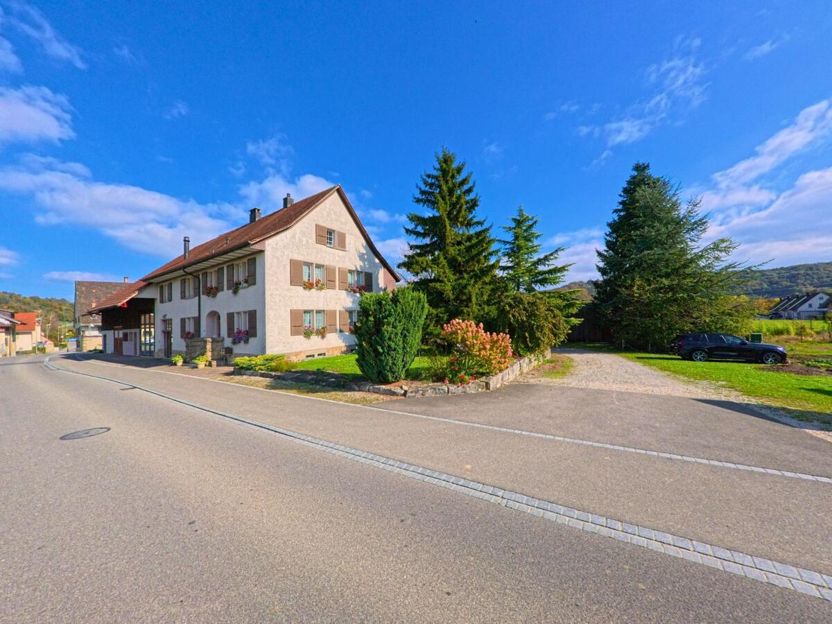 In Villnachern (5213, Kanton Aargau) steht ein traditionelles Wohnhaus mit Fensterlaeden an einer ruhig verlaufenden Strasse, waehrend hohe Nadelbaeume und eine Kieszufahrt den angrenzenden Gartenbereich praegen.