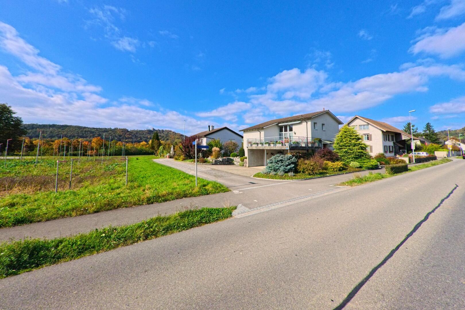 In Villnachern (5213, Kanton Aargau) reihen sich mehrere Wohnhaeuser entlang einer ruhigen Strasse, waehrend auf der linken Seite offene Felder bis zum bewaldeten Huegelrand reichen.