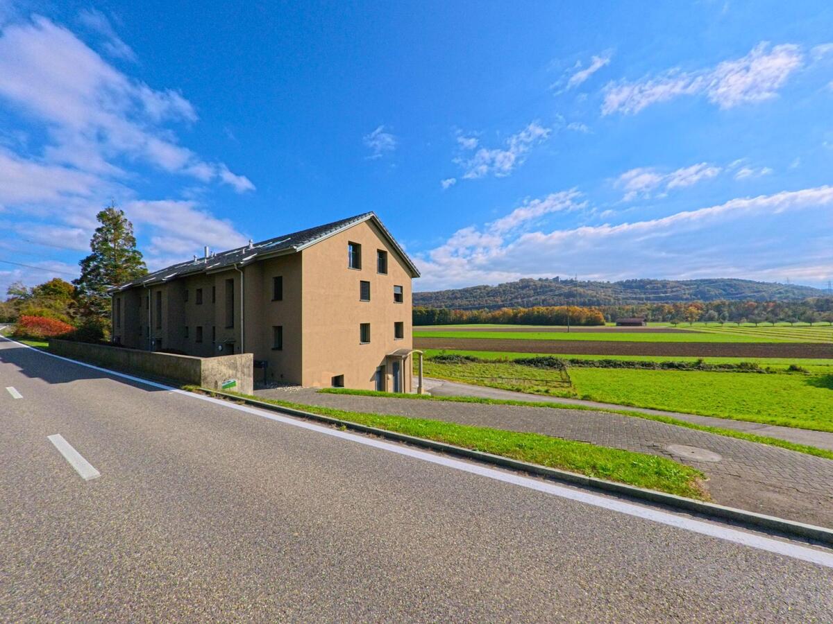 Am Ortsrand von Villnachern (5213, Kanton Aargau) steht ein modernes Wohnhaus an einer Strasse, von der aus sich der Blick weit ueber offene Felder bis zu den bewaldeten Huegeln erstreckt.