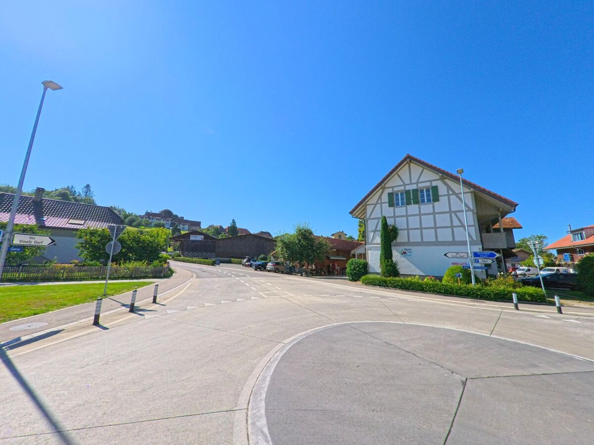 An einer Strassengabelung in Vinelz (3234, Kanton Bern) steht ein weisses Fachwerkhaus mit grünen Fensterlaeden, umgeben von Wegweisern und gepflegten Gruenflaechen.