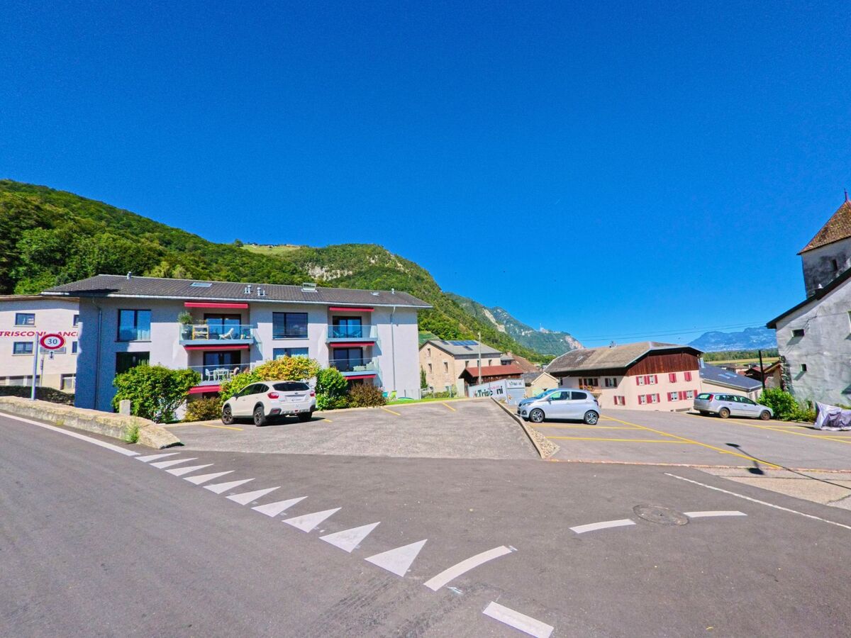 In Vionnaz (1895, Kanton Wallis) steht ein modernes Wohnhaus mit Balkonen neben einem Parkplatz, umgeben von traditionellen Gebaeuden und Bergkulisse.