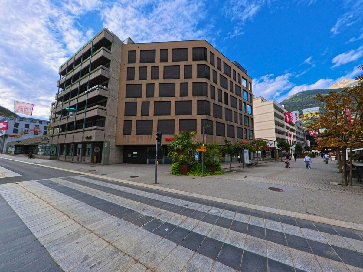 In der Innenstadt von Visp (3930, Kanton Wallis) prägen moderne Wohn- und Geschäftsbauten die Fussgängerzone, die mit Bäumen und Flaggen geschmückt ist.