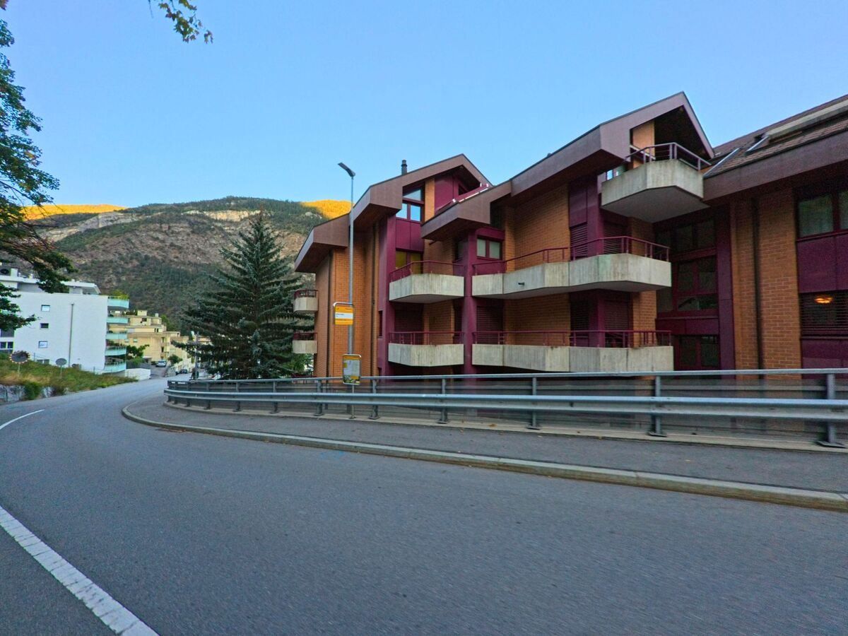 An einer Kurve in Visp (3930, Kanton Wallis) stehen Wohngebäude mit roten Balkonen, während im Hintergrund die Hänge der umliegenden Berge sichtbar sind.