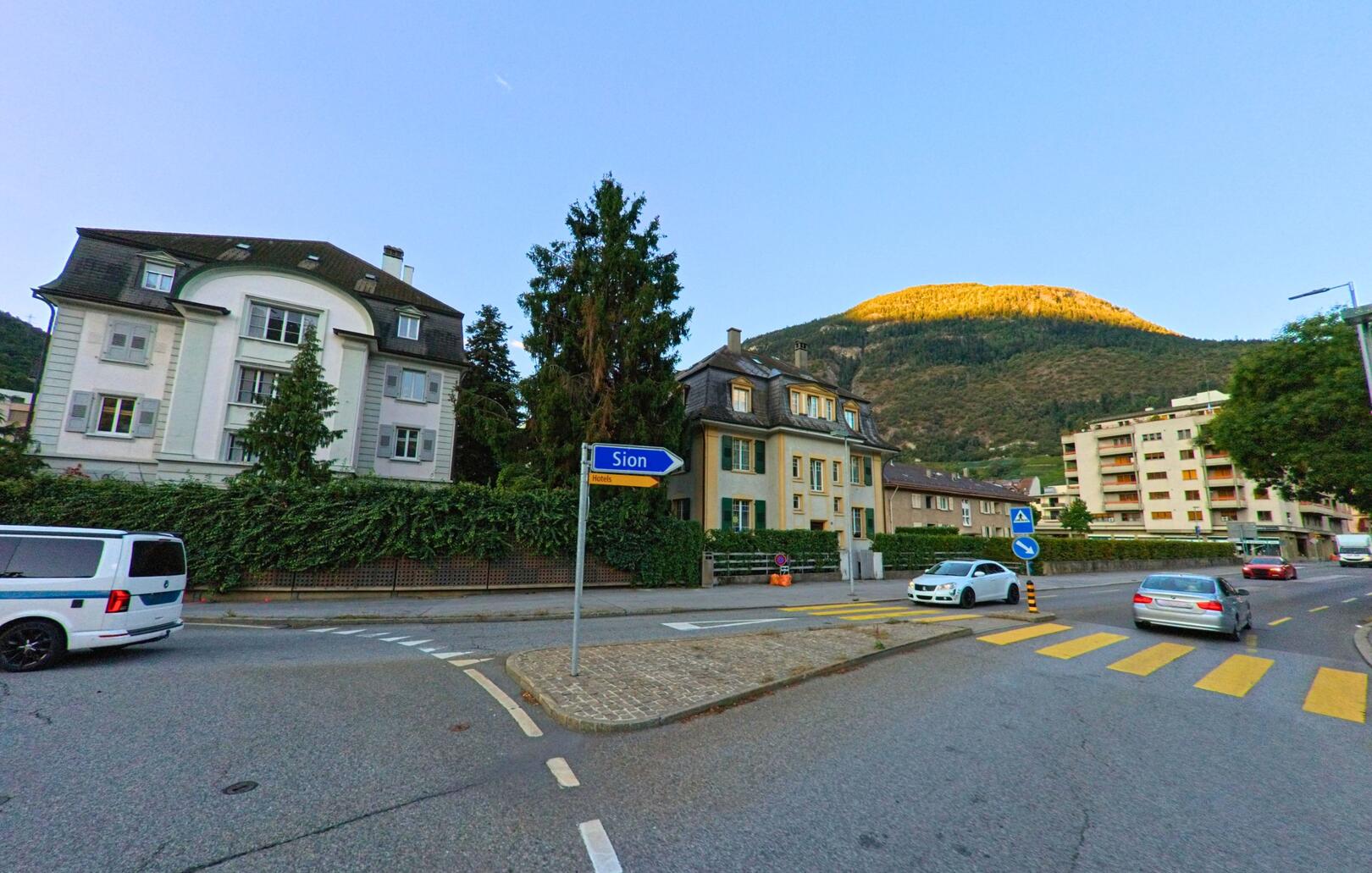 An einer Strassenkreuzung in Visp (3930, Kanton Wallis) zeigen Schilder in Richtung Sion, während im Hintergrund ein sonnenbeschienener Hügel über der Stadt liegt.