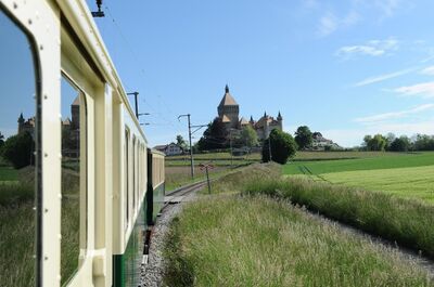Ein historischer Zug fährt durch die Felder in Morges (1110, Kanton Waadt) mit Blick auf das Schloss, das am Horizont über der Landschaft thront.