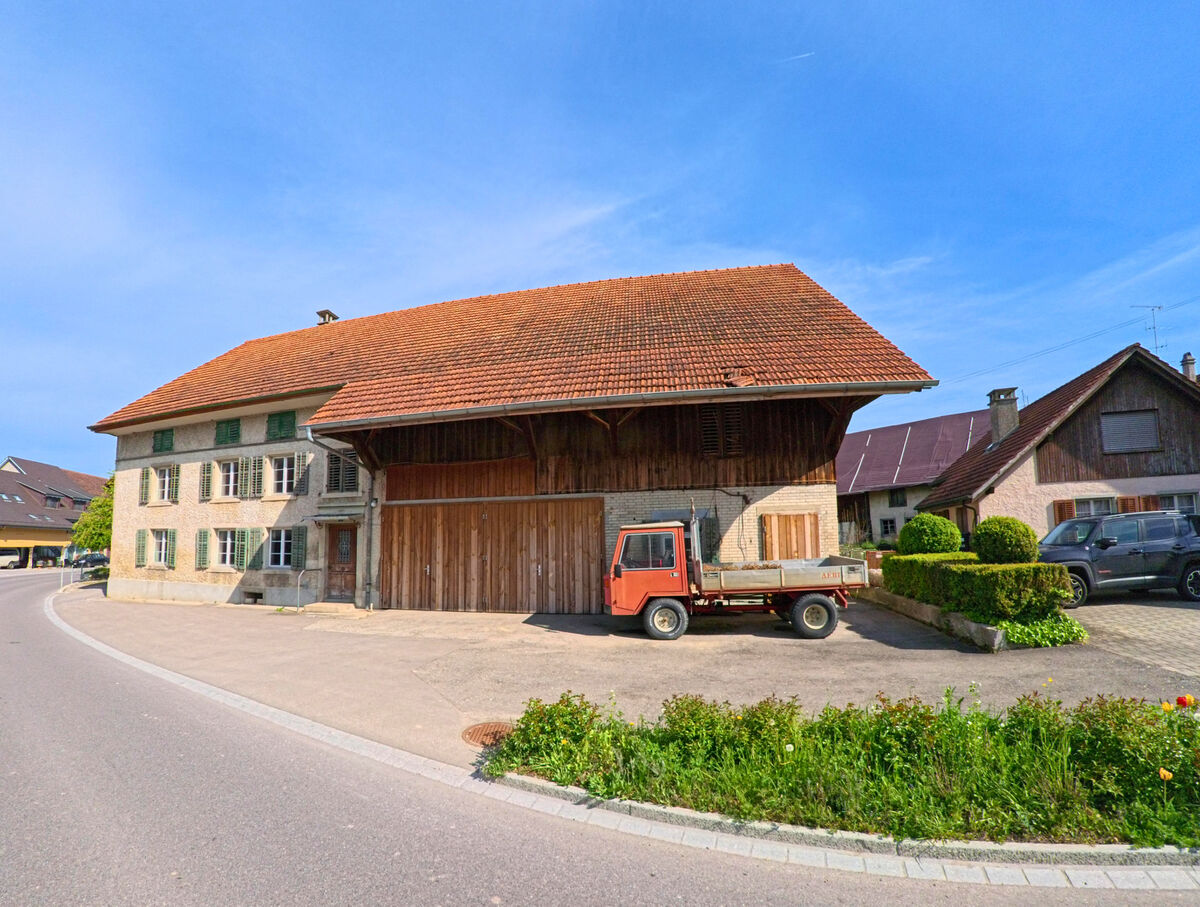 In Volken (8459, Kanton Zürich) steht ein grosses Bauernhaus mit Holzscheune und geparktem Kleintransporter an einer sanft geschwungenen Strasse.
