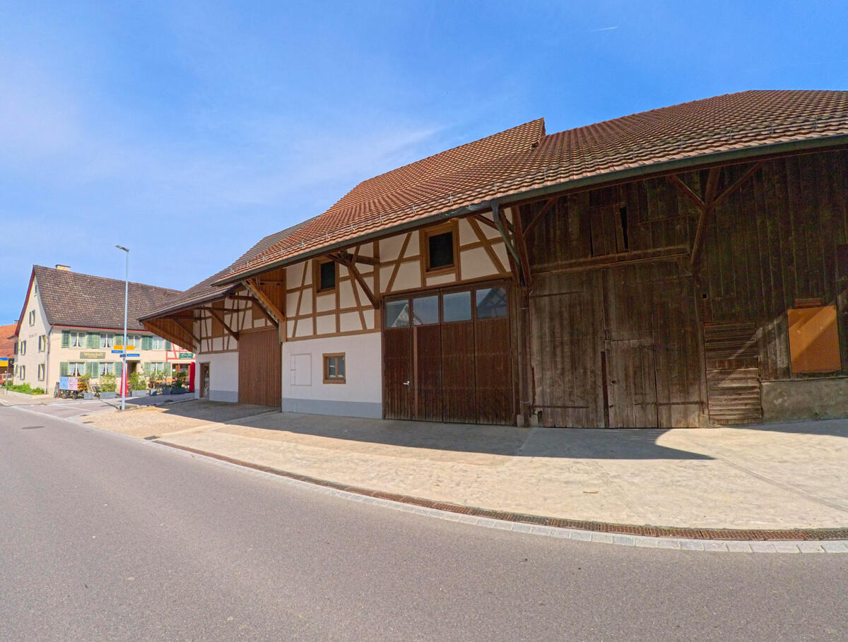 In Volken (8459, Kanton Zürich) steht eine grosse Scheune mit Fachwerkfassade und dunklem Holztor direkt an der Hauptstrasse im Dorfzentrum.