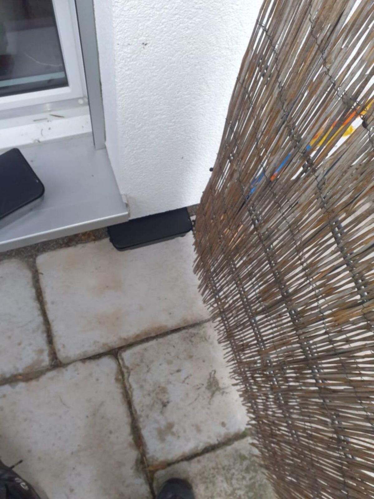 Auf einer Terrasse in Volketswil (PLZ 8604, Kanton Zürich) stehen zwei schwarze Köderboxen nahe einer Hauswand mit Fenster, teilweise verdeckt durch einen Sichtschutz aus Schilfrohr.