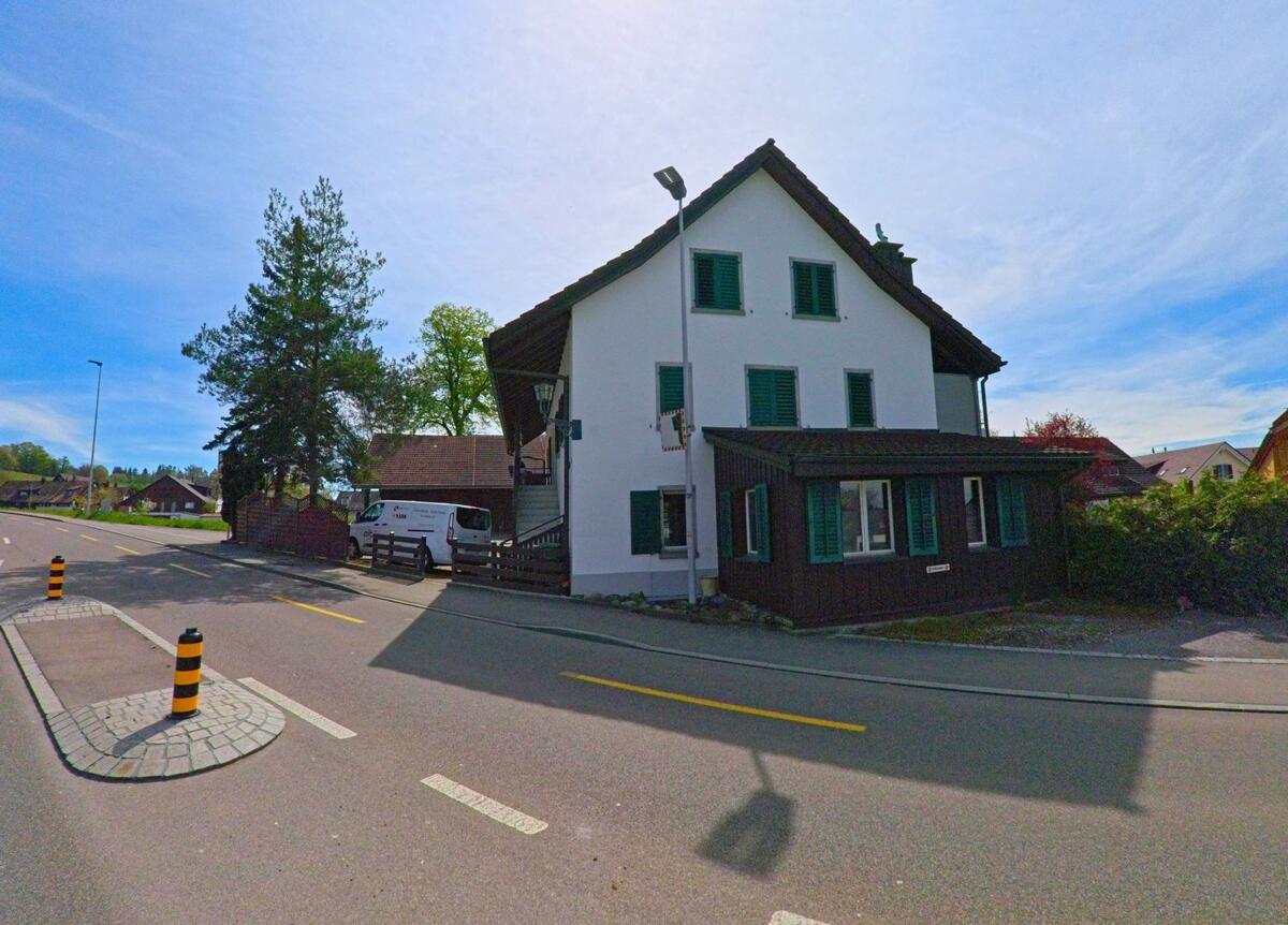 In Volketswil (8605 im Kanton Zürich) steht ein traditionelles Wohnhaus mit grünen Fensterläden an einer verkehrsberuhigten Strasse mit Sicherheitsinsel.