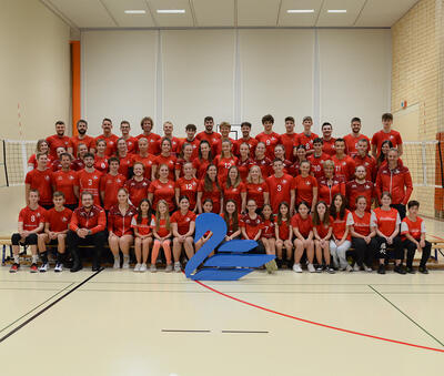 In einer Sporthalle in Bütschwil-Ganterschwil (9606, Kanton St. Gallen) posiert Volley Bütschwil mit Spielerinnen, Spielern und Nachwuchsmitgliedern in roter Vereinskleidung für ein gemeinsames Gruppenbild.