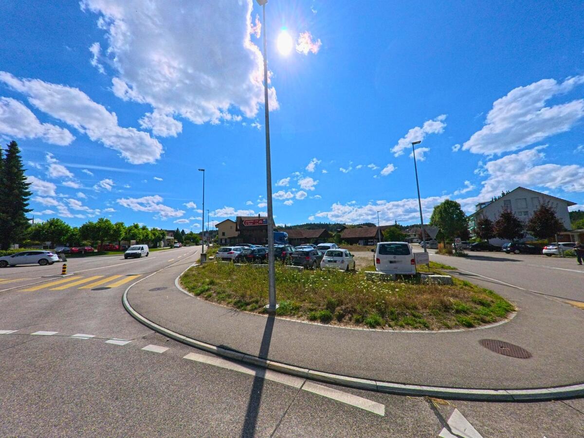 Bei Sonnenschein fuehrt eine Hauptstrasse durch Vordemwald (4803, Kanton Aargau), vorbei an einem Zebrastreifen, einem Parkplatz und verschiedenen Wohngebaeuden.