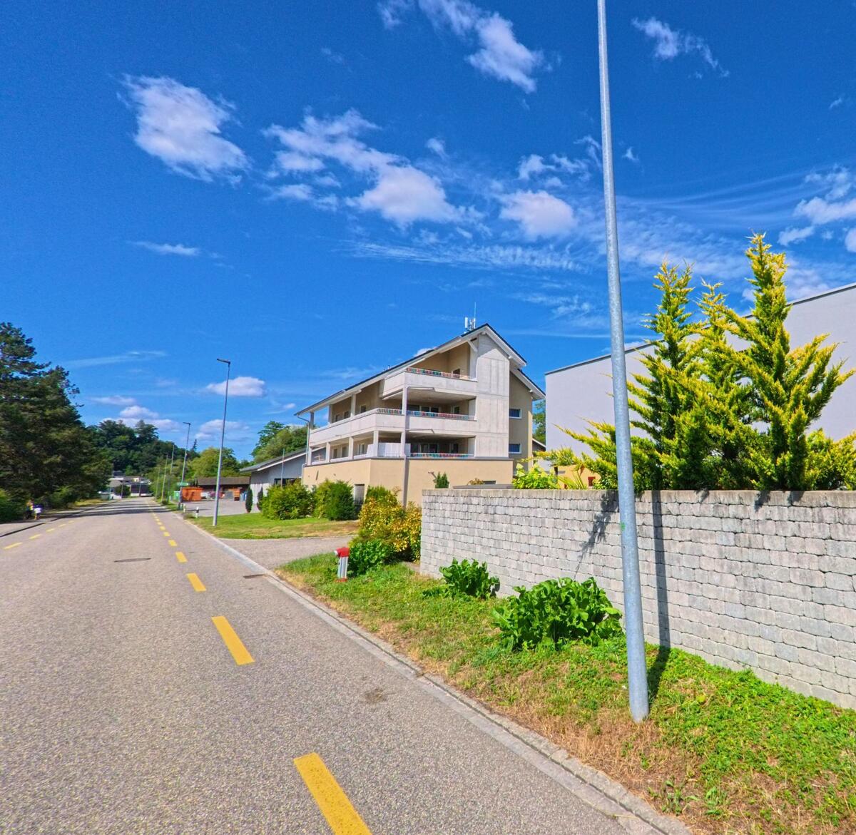 In Vordemwald (4803, Kanton Aargau) verlaeuft eine ruhige Quartierstrasse mit modernen Mehrfamilienhaeusern und gepflegten Gruenanlagen unter klarem Himmel.