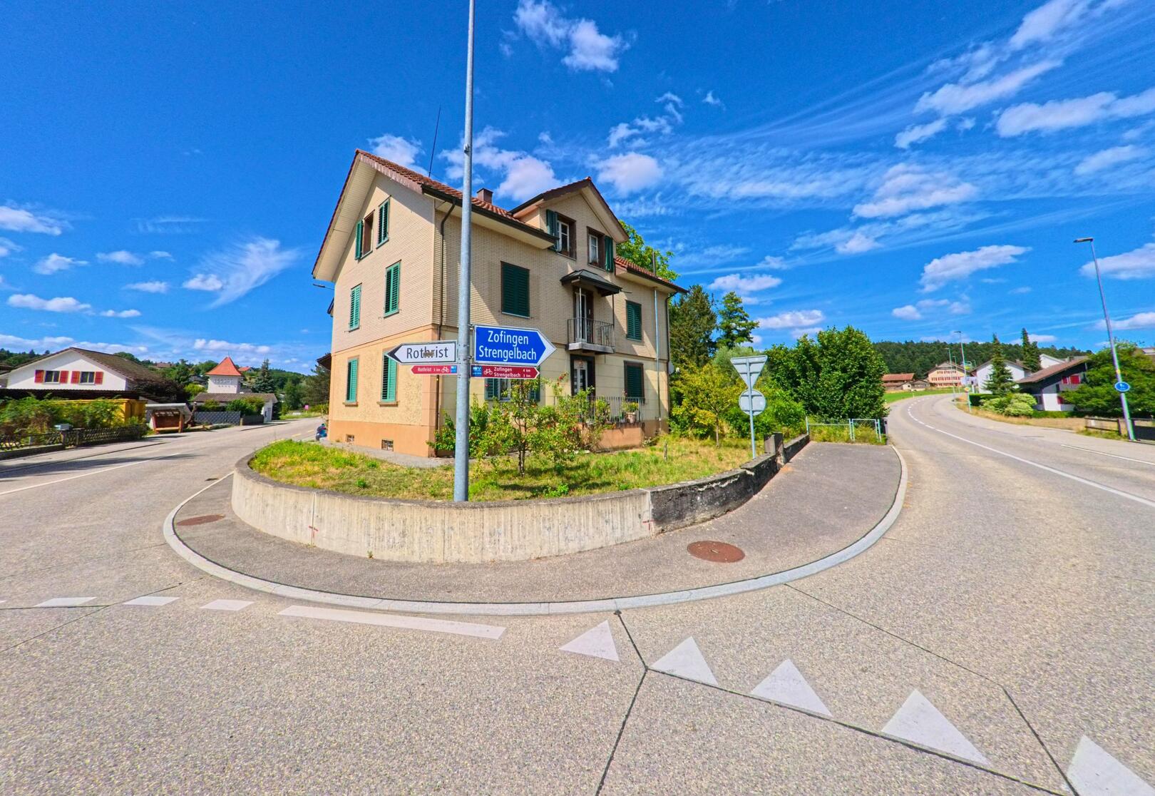 An einer markanten Weggabelung in Vordemwald (4803, Kanton Aargau) steht ein mehrstöckiges Wohnhaus mit gruenen Fensterlaeden, umgeben von Wegweisern nach Rothrist, Zofingen und Strengelbach.