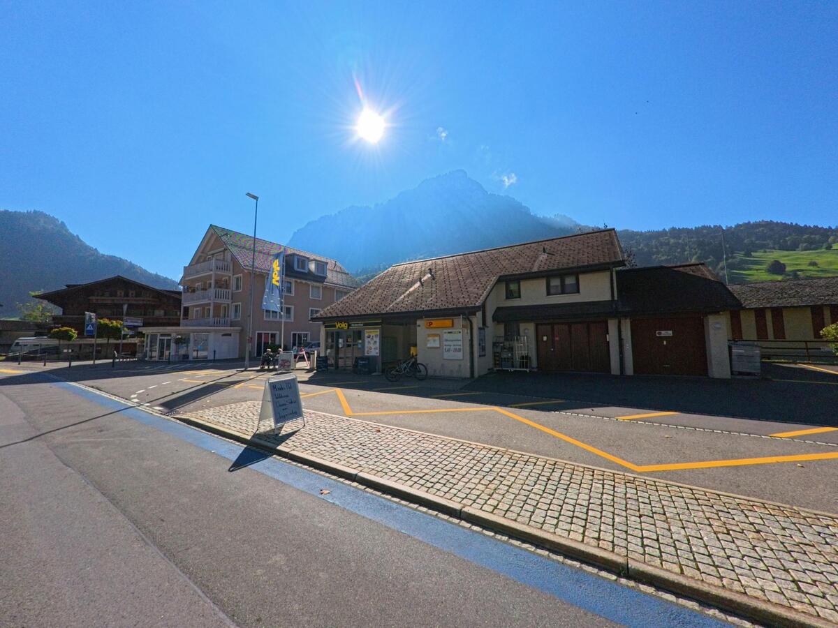 In Vorderthal (8857, Kanton Schwyz) liegt ein kleiner Dorfplatz mit Volg-Laden und weiteren Gebaeuden, waehrend die Sonne ueber den umliegenden Bergen steht.