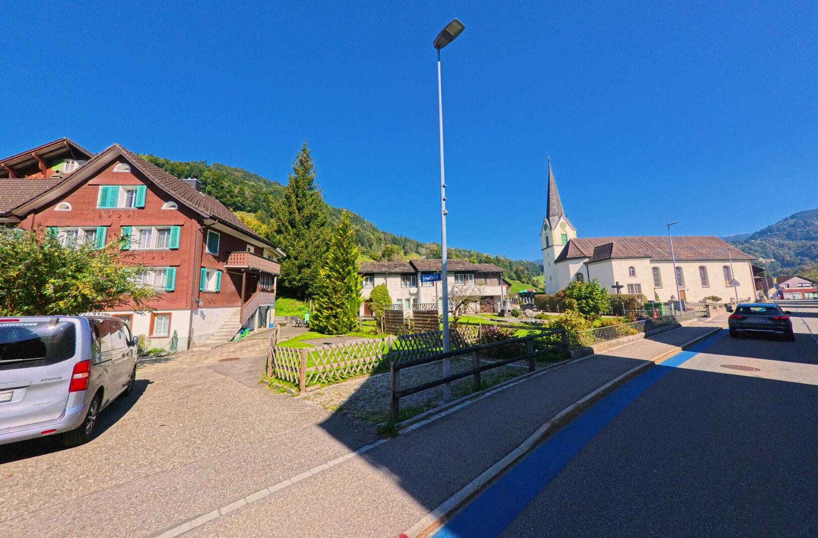 In Vorderthal (8857, Kanton Schwyz) zeigt das Bild eine Dorfstrasse mit traditionellen Haeusern und der Kirche, die vor bewaldeten Haengen deutlich hervortritt.